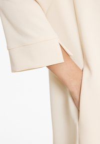 Hand in der seitlichen Tasche eines cremefarbenen, strukturierten Kleidungsstücks mit Dreiviertelarm und Manschettendetail.