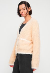 Ροδακινί χνουδωτό ζακέτα με υφασμάτινο πλεκτό σχέδιο, λευκές λεπτομέρειες κατά μήκος των άκρων και τσέπες στο μπροστινό μέρος. Κλείσιμο με κουμπιά.