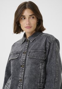 Jeansjacka i blekt svart med knappstängning framtill, två bröstfickor och krage. Har kontrasterande sömmar och en avslappnad passform.