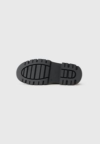 Semelle de chaussure en caoutchouc noir avec motif de grip texturé, présentant des indentations rectangulaires allongées et une zone de talon surélevée.