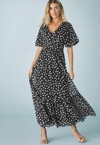 Musta pilkullinen maxi-mekko, jossa on kietaisupääntie, lyhyet puffihihat ja röyhelöhelma. Mekko on valmistettu kevyestä kankaasta ja siinä on ilmava muotoilu.