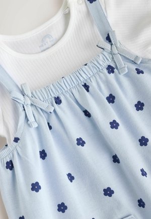 Blaues Baumwollkleid mit marineblauen Blumenmustern und verstellbaren Schulterriemen, getragen über einem weißen, gerippten Langarmshirt.