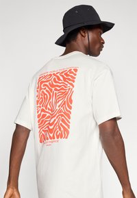 Λευκή βαμβακερή μπλούζα με large πορτοκαλί αφηρημένο σχέδιο στην πλάτη, με κείμενο "FREERIDE WORLD TOUR" και "HOME OF FREERIDE" εκτυπωμένα από κάτω.