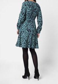 Teal kukka-asusteinen mekko, jossa on pitkät hihat, elastinen vyötärö ja leveä helma. Tumma kukkakuosi ja pleatattu takakuvio. Yhdistetty mustien sukkahousujen ja nilkkureiden kanssa.