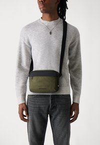 Grüne und schwarze Umhängetasche mit glatter Textur, rechteckiger Form und Reißverschlussverschluss, getragen über einem grauen Pullover und dunklen Jeans.