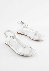 Silverfärgade metalliska sandaler med justerbara ankelremmar, platta sulor, öppen tå och en minimalistisk design med korsremmar och en slät yta.