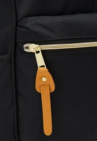 Czarny materiałowy plecak z złotym zamkiem błyskawicznym i brązowym zamszowym uchwytem zamka. Powierzchnia jest teksturowana i trwała.