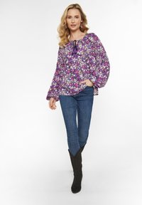 Lila Bluse mit floralen Mustern und langen Ballonärmeln, mit einer Schleife am Ausschnitt. Kombiniert mit blauen Jeans und schwarzen Stiefeletten.