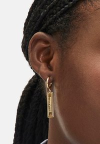 Gouden creolen met een rechthoekige hanger met gegraveerde tekst. Het ontwerp heeft een gestructureerde afwerking en een glanzend oppervlak.