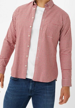 Mand iført en rød og hvid småternet skjorte over en hvid t-shirt sammen med blå jeans, stående mod en ensfarvet baggrund.