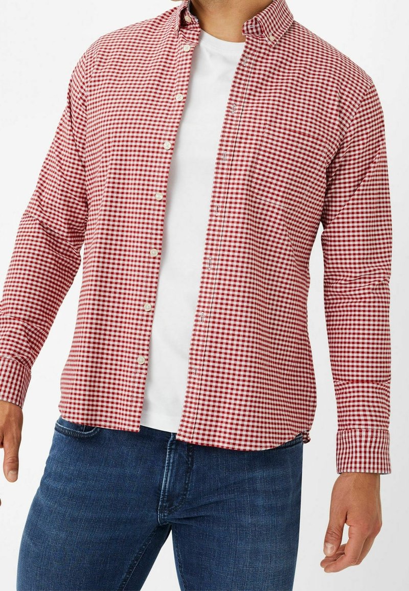 Homme portant une chemise à petits carreaux rouge et blanc boutonnée sur un t-shirt blanc, associée à un jean bleu, debout devant un fond uni.