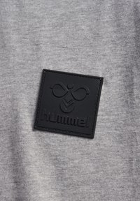Gråt stof med en blød tekstur har en sort gummipatch med et logo og navnet "hummel" prægede i midten.