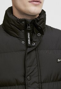 Schwarze gepolsterte Jacke mit hohem Kragen, Druckknöpfen und Frontreißverschluss. Sie verfügt über ein strukturiertes Material und ein Logo-Detail auf der Brust.