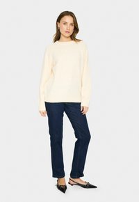 Kvinna i en krämfärgad långärmad tröja, mörkblå jeans och svarta spetsiga slingback-sandaler, stående mot en enkel ljus bakgrund.
