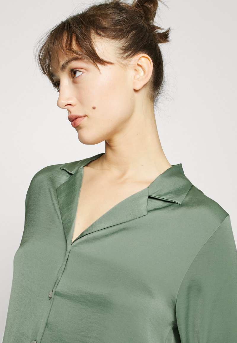Camicia di raso verde con colletto, caratterizzata da una chiusura con bottoni sul davanti e una texture liscia e lucida. Il design ha una vestibilità rilassata e moderna.