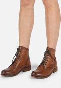 Botas de cuero marrón hasta el tobillo con punteras redondeadas, cordones en la parte delantera y tacones de madera bajos. Textura suave con detalles de costura en los laterales.