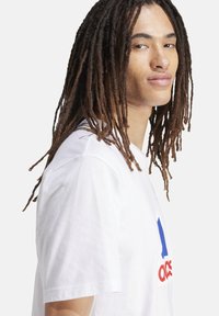 T-shirt bianco in cotone con un grande logo grafico blu e rosso sul davanti, con scollo rotondo e maniche corte.