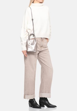 Persona che indossa un maglione a collo alto color crema, pantaloni beige a gamba larga risvoltati alle caviglie, stivali neri con punta e una piccola borsa metallica con tracolla a catena.