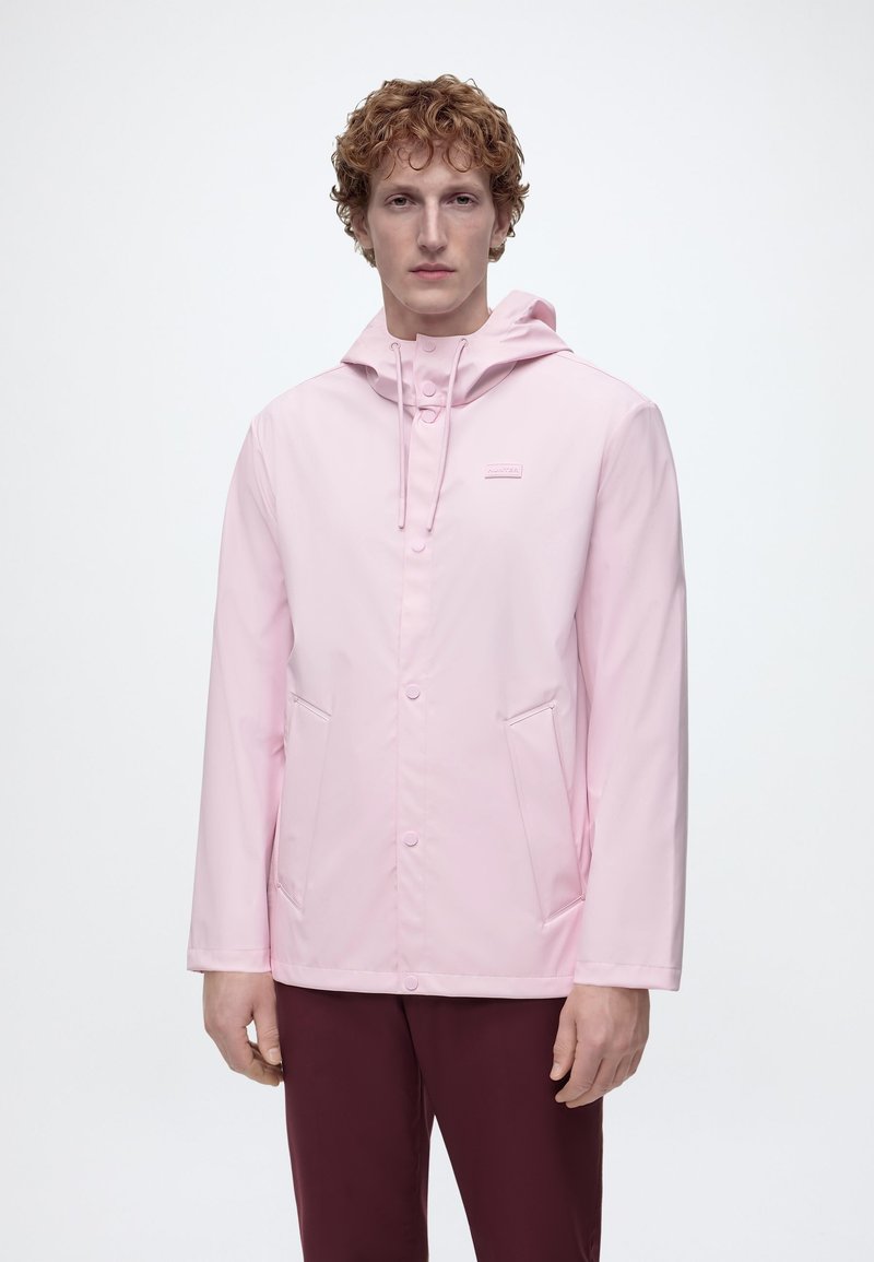 Άνδρας με σγουρά κόκκινα μαλλιά, φοράει ανοιχτό ροζ αδιάβροχο με κουκούλα και σκούρο μαρόν παντελόνι, μπροστά σε ένα απλό λευκό φόντο.