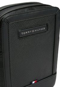 Bolso de cuero negro con una textura de grano, que presenta una placa de logo rectangular y un acento tricolor en la parte inferior. Cierre de cremallera.