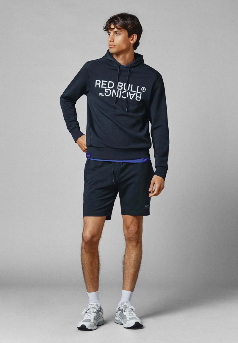 Sudadera azul marino con el logo blanco "RED BULL RACING", combinada con pantalones cortos negros y zapatillas grises. Capucha, cordones y detalle interior morado.