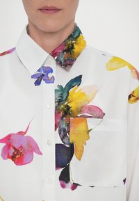 White floral shirt featuring a multi-colored print with vibrant flowers. Includes a patch pocket and a classic collar with contrasting accents.