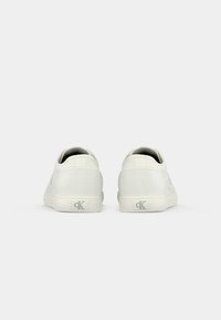 Para białych niskich sneakersów pokazana od tyłu z logo "cK" na pięcie, na jednolitym jasnoszarym tle.