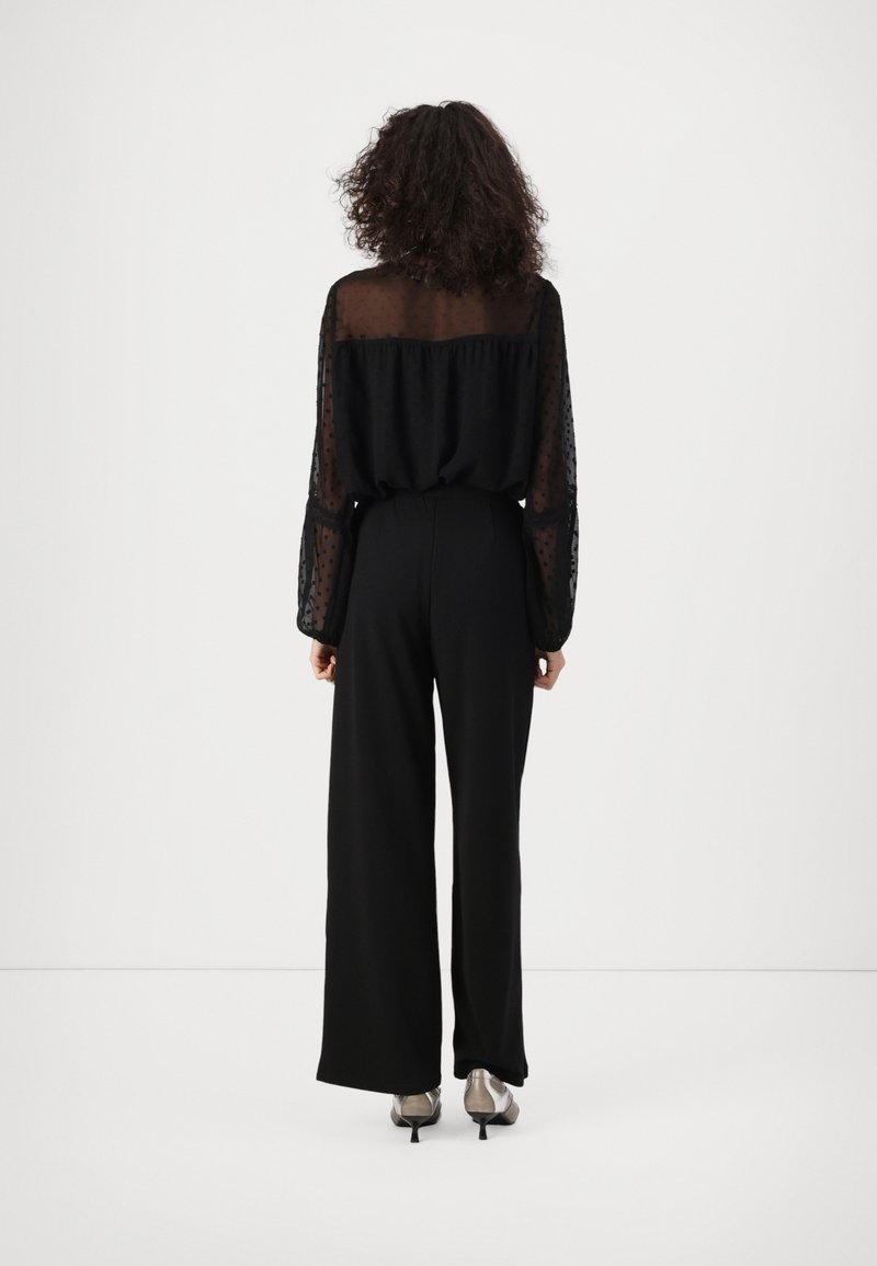 Blusa nera a maniche lunghe in tessuto trasparente con un motivo a pois, abbinata a pantaloni neri a vita alta. Il modello indossa scarpe con tacco metalliche.