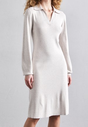 Kobieta w jasnobeżowej dzianinowej sukience z długim rękawem sięgającej do kolan, z kołnierzem i dekoltem w serek, stojąca na tle jednolitego szarego tła.