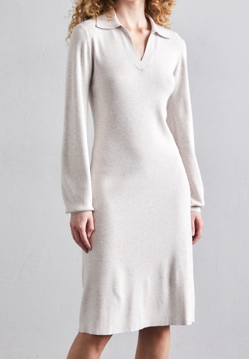 Femme portant une robe en maille beige clair à manches longues, longueur genou, avec col et encolure en V, debout devant un fond gris uni.