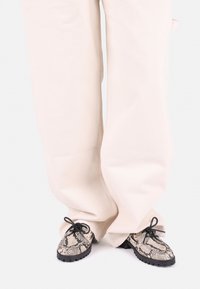 Laced shoes with a black rubber sole, featuring a snakeskin pattern in grey and beige. Worn with loose-fitting, cream-colored pants.