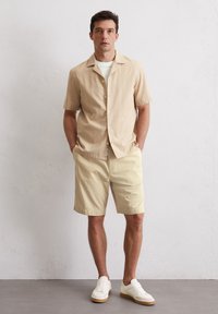 Lichtbeige korte mouw shirt met knoopsluiting, gecombineerd met lichtgele shorts. Witte sneakers met een gumzool maken de outfit compleet.