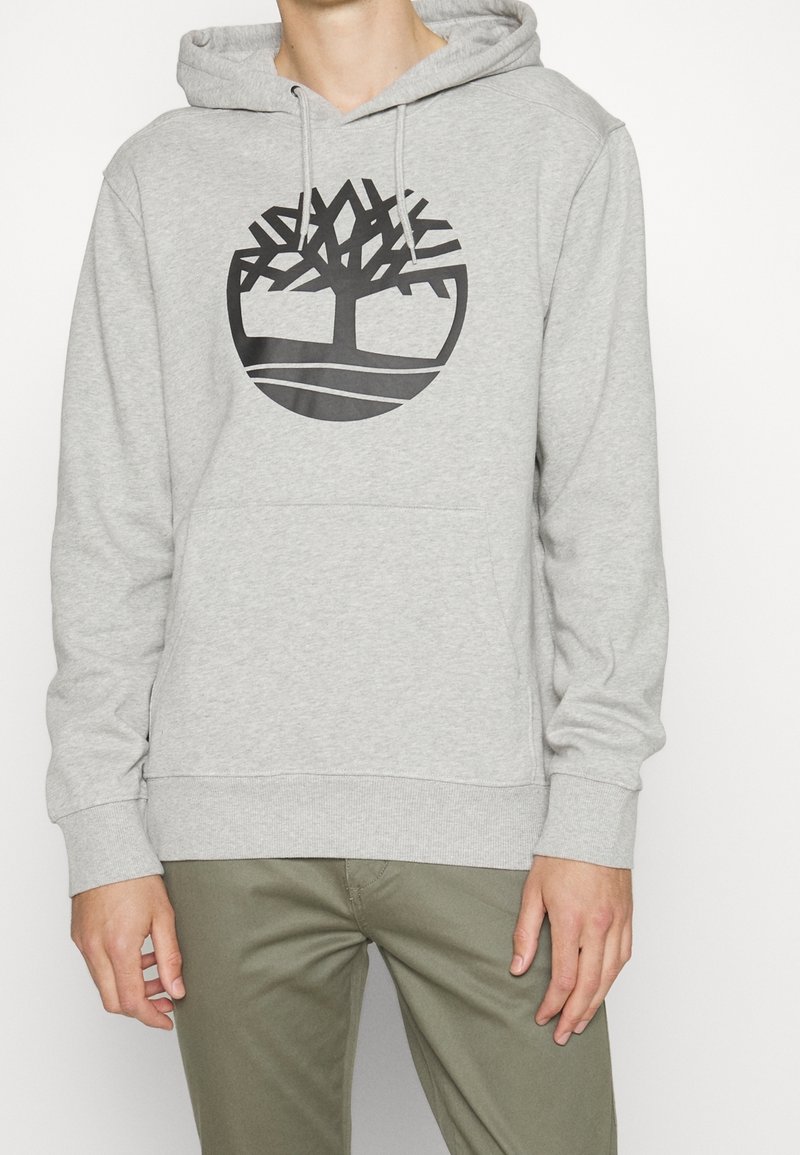 Personne portant un sweat à capuche gris clair avec un logo d'arbre stylisé noir, associé à un pantalon vert olive, debout sur un fond uni.