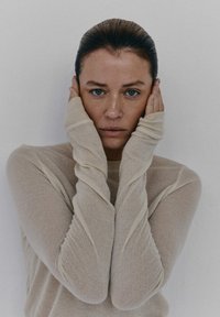 Beżowa, długi rękaw bluzka z miękkiego, teksturowanego materiału, z dopasowanymi mankietami. Osoba trzyma twarz obiema rękami, na neutralnym tle.