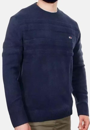 Uomo con un maglione blu scuro a maglia con un piccolo logo rettangolare rosso, bianco e blu sul petto sinistro, abbinato a pantaloni marrone chiaro.