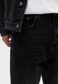 Gros plan sur une personne portant un jean en denim noir et une veste en denim noir assortie avec des détails de boutons métalliques.