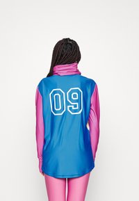 T-shirt de sport à manches longues avec un dos bleu présentant le numéro blanc "09", des manches roses, un col haut et un tissu lisse et léger.