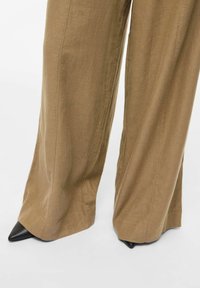 Pantalon beige à jambes larges couvrant des chaussures noires à bouts pointus, sur fond blanc.