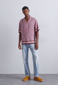 Chemise à manches courtes bourgogne avec des motifs géométriques, associée à un jean bleu clair et des baskets jaunes. Col simple et coupe décontractée.