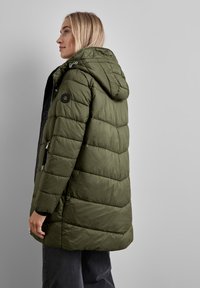 Olivgrön pufferjacka med huva, som har horisontell quiltad textur, sidofickor med dragkedja, svarta detaljer och en logotyp på ärmen.