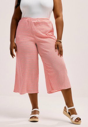 Donna che indossa pantaloni larghi a righe rosse e bianche, tagliati sopra la caviglia, top bianco senza maniche e sandali con zeppa bianchi con cinturini alla caviglia.