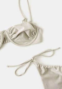 Ensemble bikini beige clair avec haut à armatures froncées et bas à nouer sur les côtés, posé à plat sur une surface blanche.