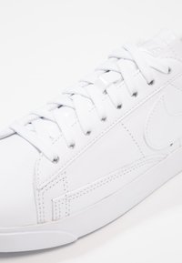 Zapatilla de cuero blanca con textura suave, diseño con cordones y acentos de marca. Tiene un perfil bajo y suela de goma.