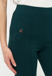 Gros plan sur un pantalon ajusté vert foncé avec un petit rabat de poche et un bouton rond doré près de la taille, porté avec un haut blanc.