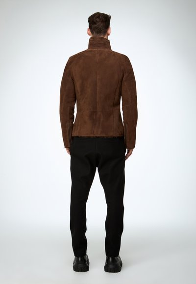 Braune Wildlederjacke mit hohem Kragen, tailliertem Schnitt und seitlichen Reißverschlüssen. Kombiniert mit schwarzer Hose und klobigen schwarzen Schuhen.