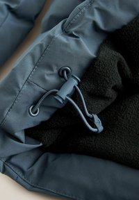 Detaliu al unui jacheta soft-shell, cu un strat exterior albastru, o căptușeală din fleece negru și un sistem de ajustare cu șnur și toggles din plastic.