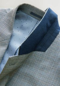 Textureret brun og blå jakke krave med en blå mønstret foer, der har et rutenet mønster og fine prikkede detaljer.