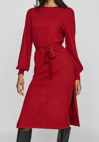 Czerwona sukienka z długim rękawem i paskiem w talii, o luźnym kroju i bocznym rozcięciu. Wykonana z miękkiego materiału o delikatnej teksturze.