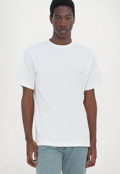 T-shirt en coton blanc avec un col rond et des manches courtes, présentant un petit accent brodé sur le côté gauche de la poitrine. Associé à un jean bleu clair.