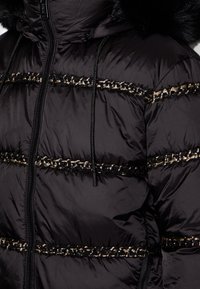 Veste matelassée noire avec garniture en chaîne dorée et noire sur la poitrine et les manches, fermeture éclair à l'avant, cordons de serrage et capuche doublée de fourrure.